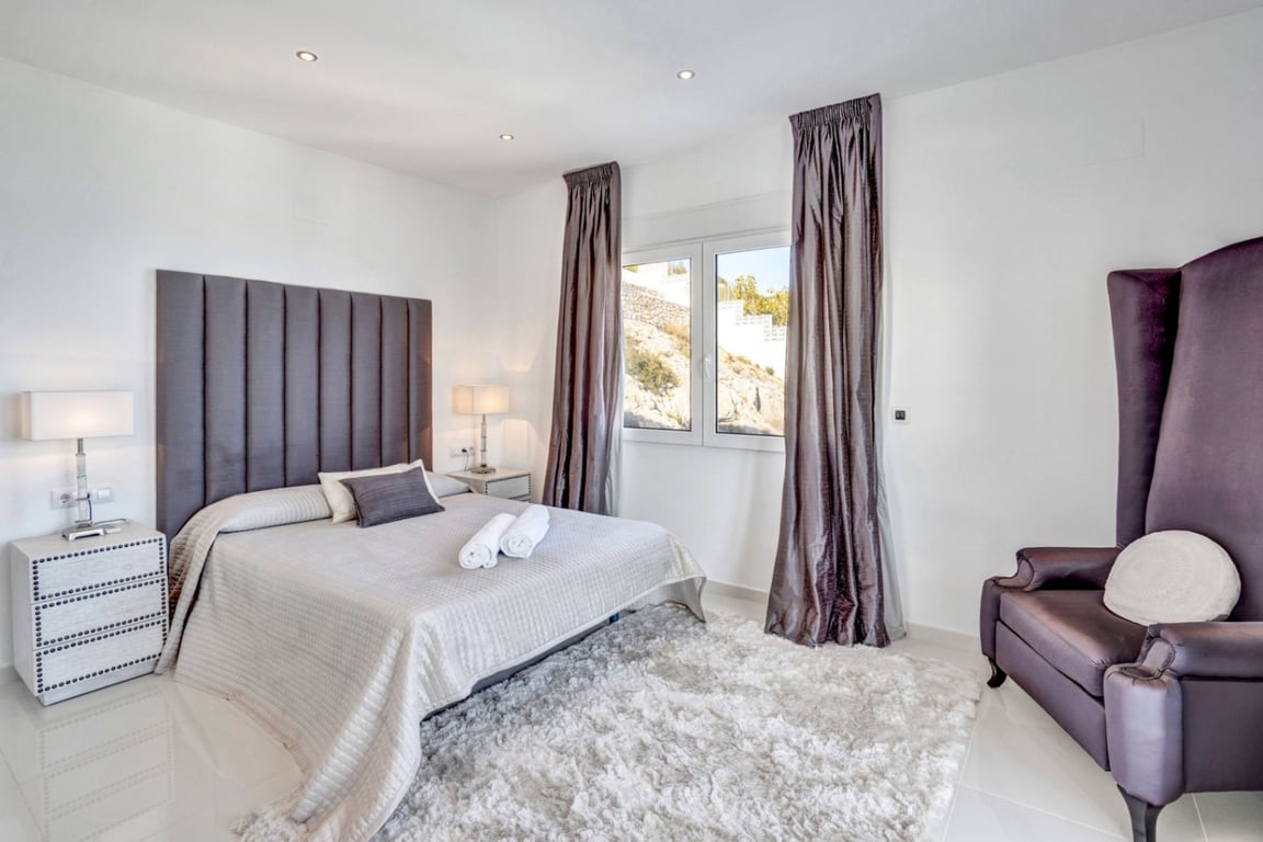 Dormitorio con cama doble, cabecero tapizado en gris y sillón de respaldo alto. Incluye alfombra de pelo largo, iluminación empotrada y ventana con vistas a la montaña.