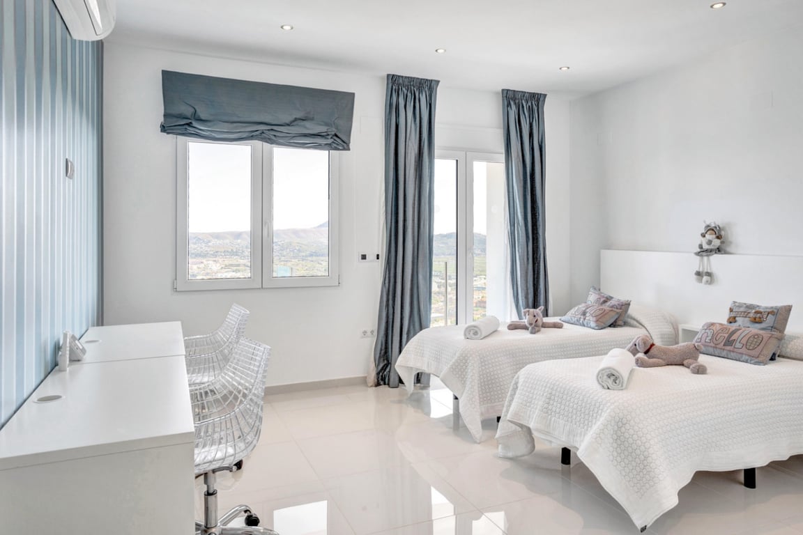 Dormitorio con dos camas individuales, escritorio blanco con sillas transparentes y ventanales con vistas panorámicas a la montaña y el mar.