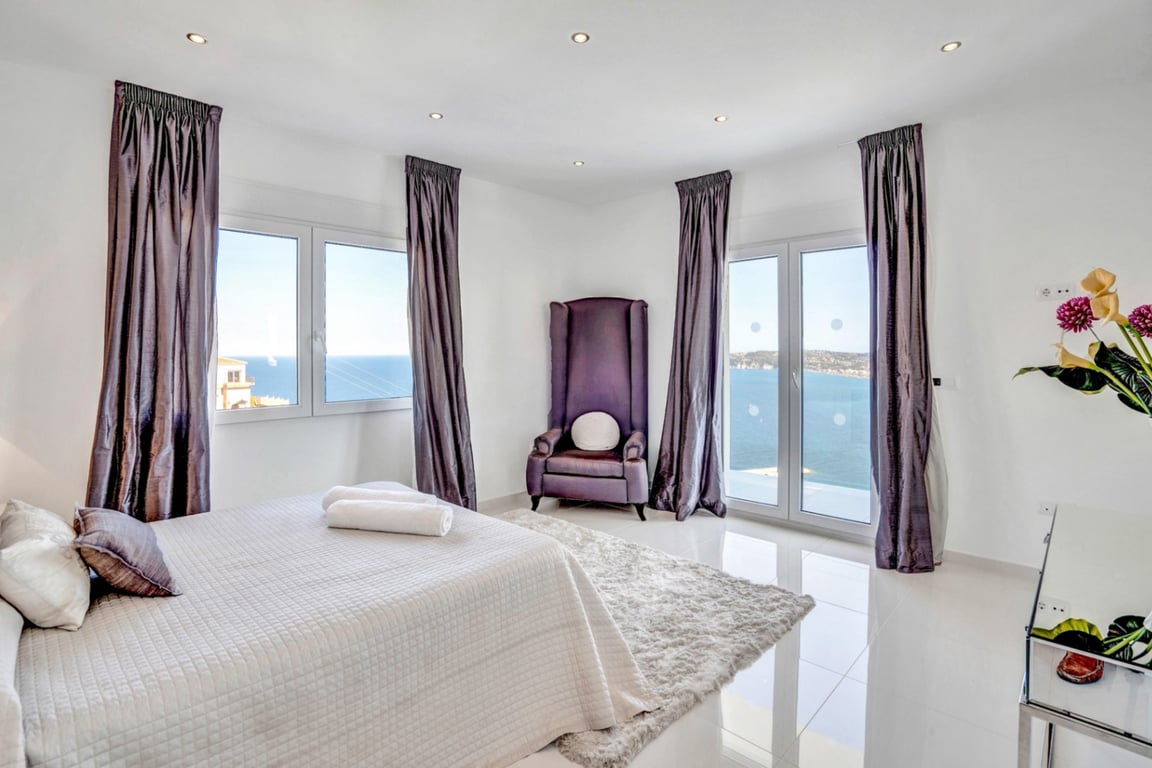 Dormitorio principal con cama doble, sillón de respaldo alto en terciopelo violeta y ventanales con vistas panorámicas al mar Mediterráneo.
