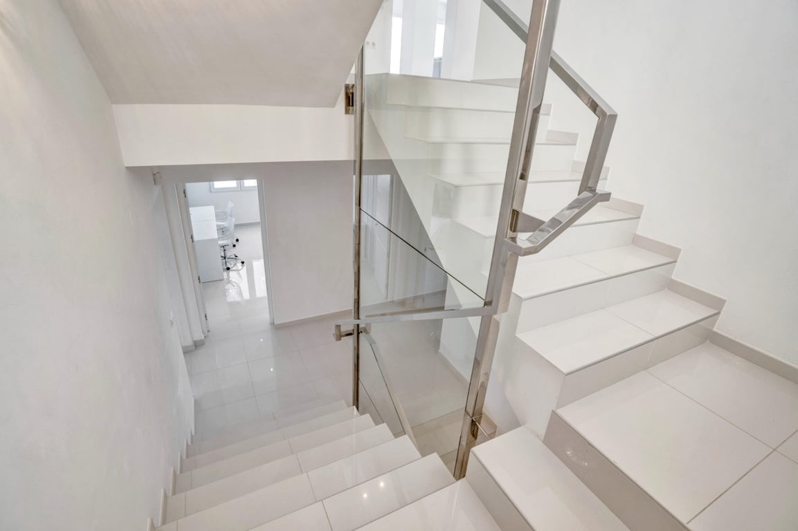 Escalera moderna con peldaños blancos, barandilla de cristal y acero inoxidable que conecta los niveles de la villa con un diseño limpio y luminoso.