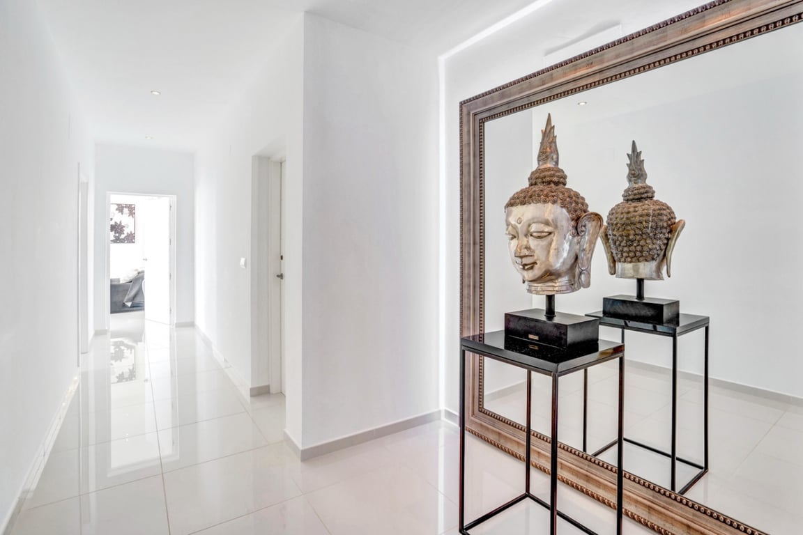 Pasillo minimalista con suelo de gres pulido e iluminación empotrada. Destaca un gran espejo decorativo y una escultura de Buda sobre un pedestal negro.