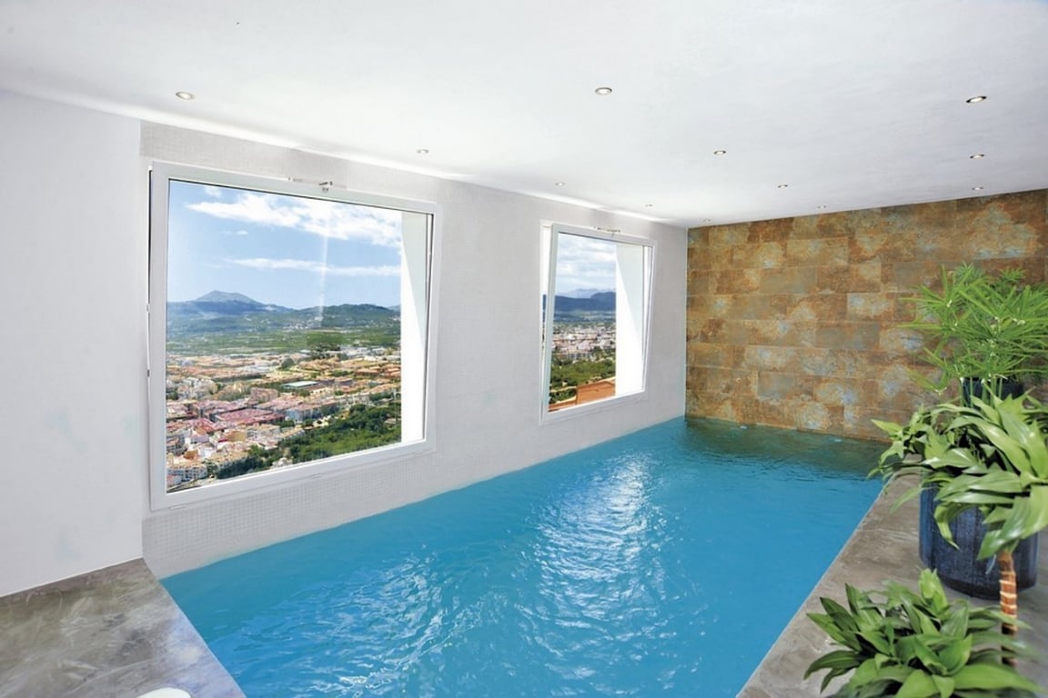 Piscina interior climatizada con ventanales panorámicos hacia el valle y la montaña, destacando una pared de piedra natural y focos empotrados en el techo.