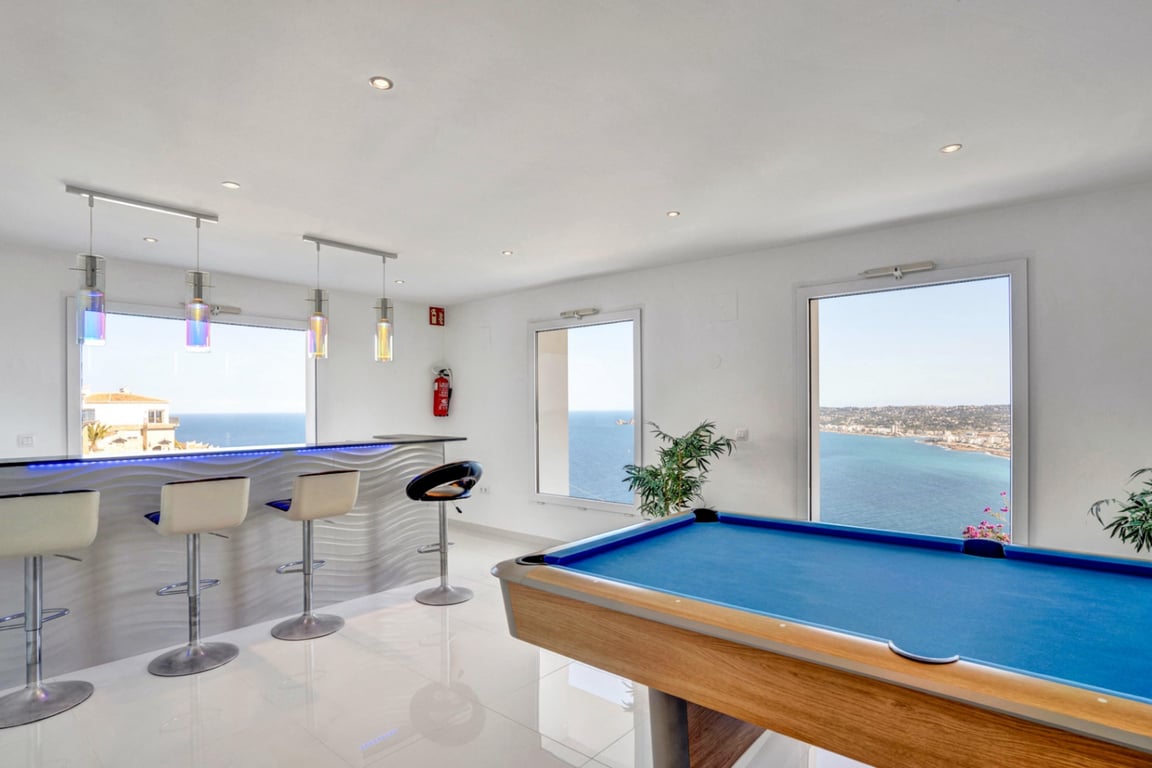 Sala de ocio con mesa de billar azul y bar moderno con iluminación LED, ofreciendo vistas panorámicas al mar a través de sus amplios ventanales.