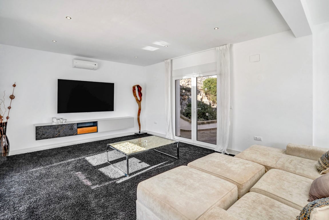 Sala multimedia moderna con sofá modular, alfombra de pelo largo y mueble flotante, con salida directa a la terraza integrada en la roca natural.