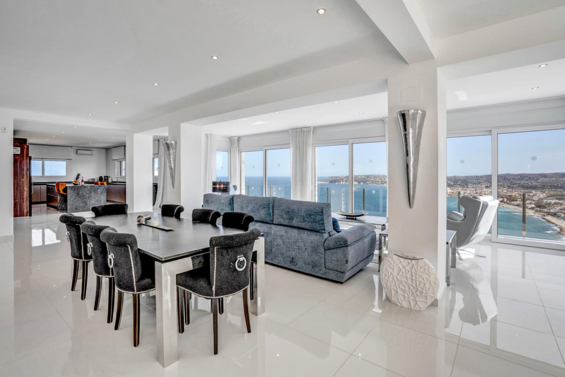 Salón-comedor de planta abierta con suelos pulidos, mobiliario moderno y amplios ventanales que ofrecen vistas panorámicas al mar y a la bahía de Jávea.