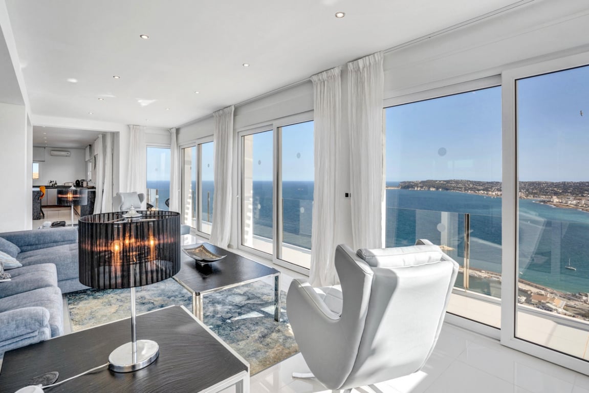 Salón luminoso con ventanales correderos de suelo a techo y vistas panorámicas al puerto de Jávea. Dispone de mobiliario moderno, suelos cerámicos claros y focos empotrados.