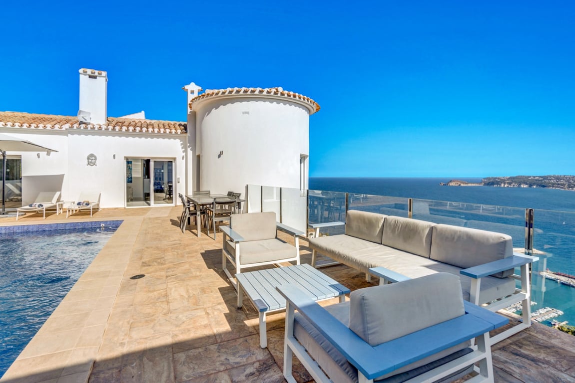 Terraza principal con zona de estar, comedor exterior y piscina, ofreciendo vistas panorámicas al puerto de Jávea y al mar Mediterráneo tras una barandilla de cristal.