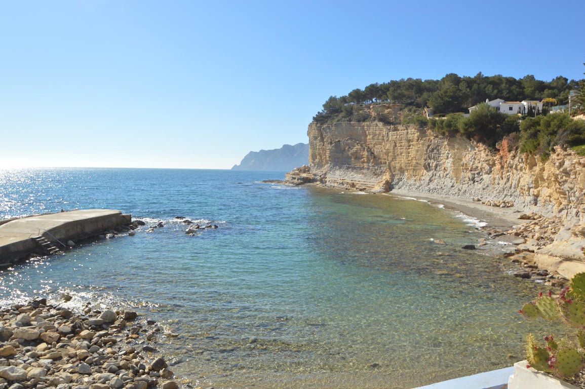 Vista de Cala Advocat con aguas cristalinas, un pequeño espigón de hormigón y acantilados de piedra caliza bajo un cielo despejado en la costa de Benissa.