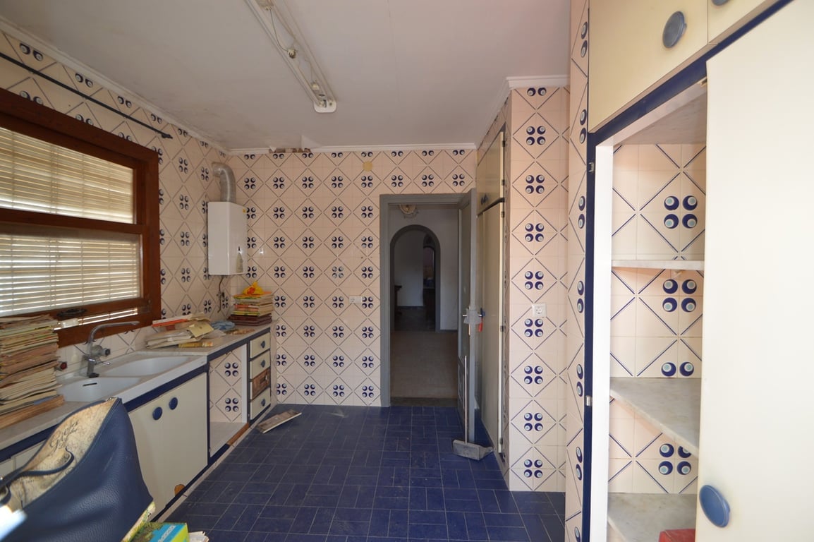Cocina con azulejos tradicionales, suelos azules y armarios blancos con bordes azules. Dispone de fregadero doble bajo ventana de madera y conexión al pasillo central.