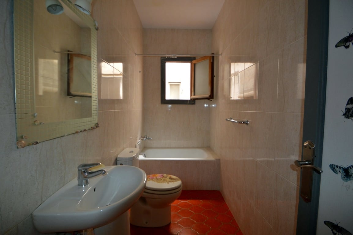 Cuarto de baño con suelos cerámicos rojos hexagonales, paredes alicatadas en tonos crema, lavabo de pedestal y una bañera encastrada situada bajo una ventana exterior.