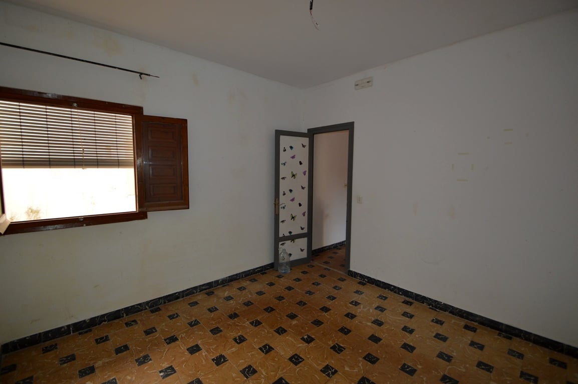 Dormitorio con suelos de baldosa tradicional, paredes blancas y ventana de madera con contraventanas. Una estancia con gran potencial de reforma.