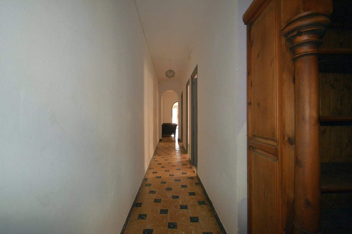 Pasillo alargado con suelos de baldosa decorada y paredes blancas, que conduce a un arco con luz natural y un mueble de madera tallada en primer plano.