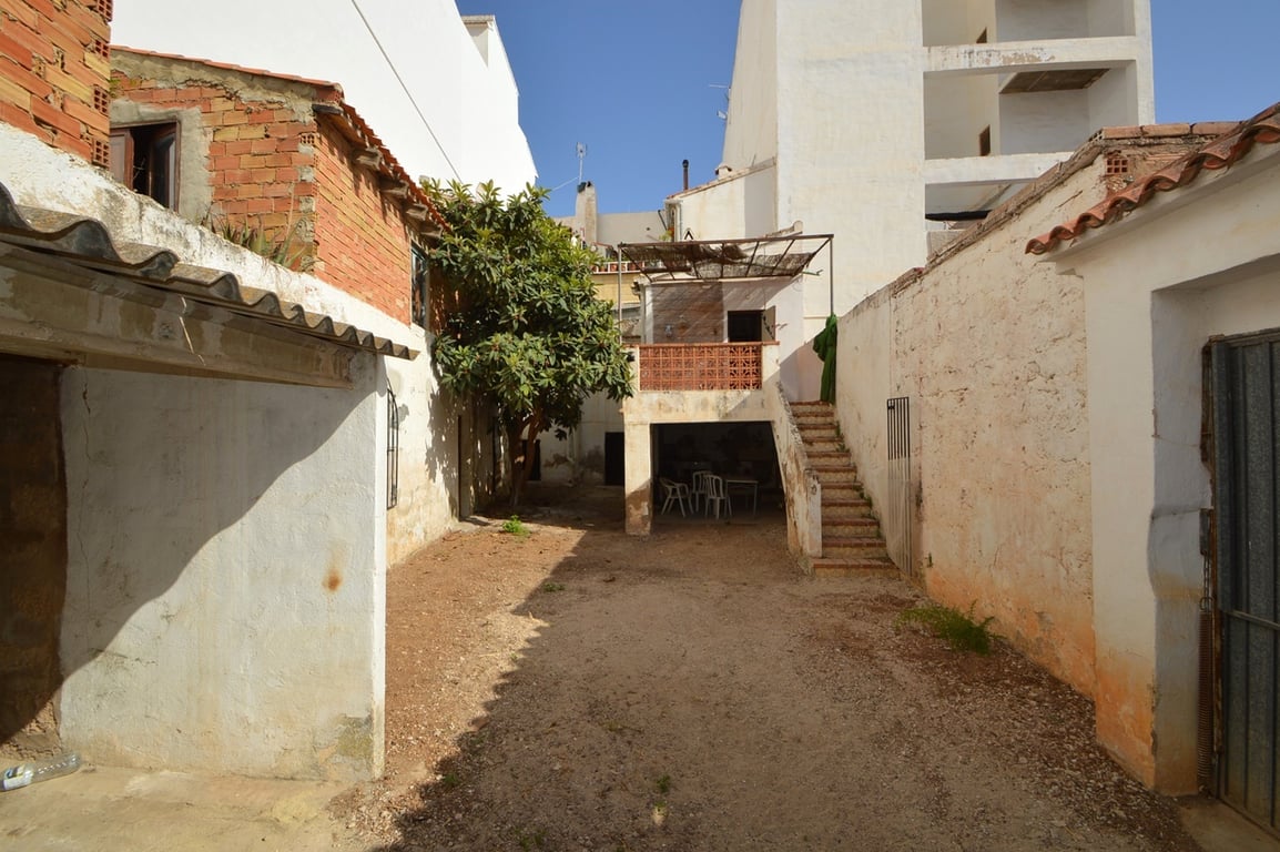 Patio interior privado con suelo de tierra, muros altos y una escalera exterior que conduce a una terraza elevada. Incluye una zona cubierta y un árbol maduro.