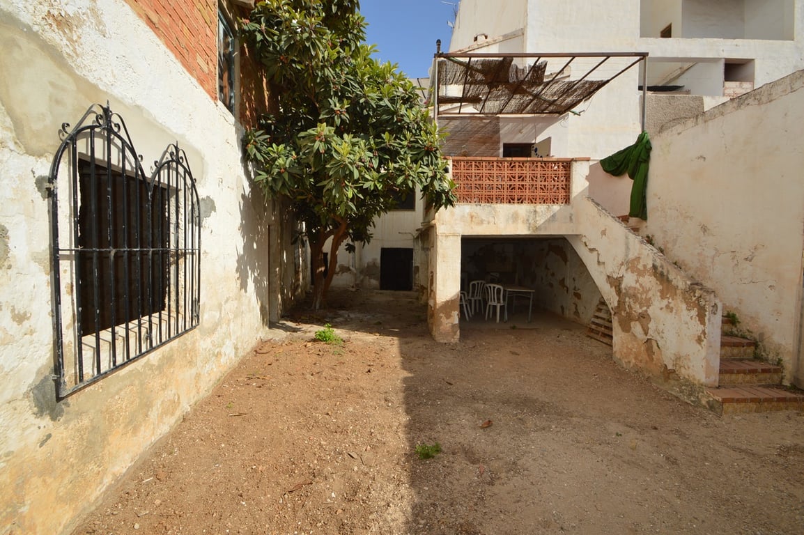 Patio privado de tierra con un árbol maduro, ventana con rejería de hierro y acceso a una terraza superior mediante escaleras exteriores de piedra.