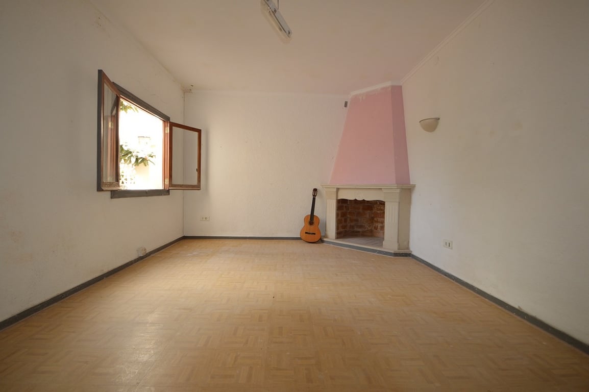 Salón diáfano con chimenea de rincón en tono rosa, suelos de parqué y ventana de madera con vistas al exterior. Un espacio versátil con luz natural.