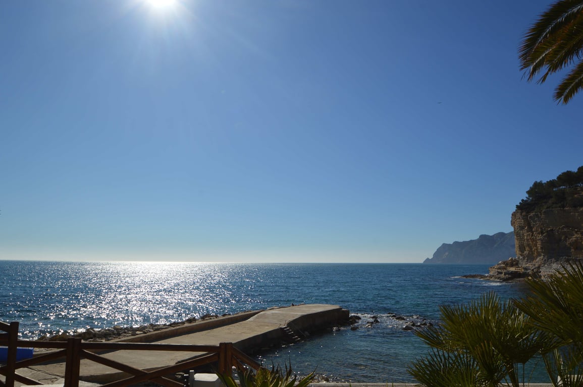 Vista panorámica desde la costa de Benissa hacia el mar Mediterráneo, destacando un pequeño muelle de hormigón y los acantilados bajo un cielo despejado.