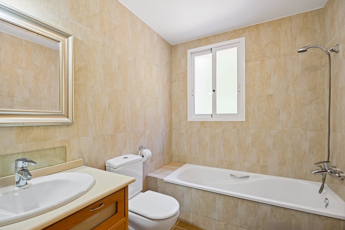 Baño funcional con paredes alicatadas en tonos beige, bañera integrada con ducha, lavabo con mueble de madera y ventana de climalit para luz natural.