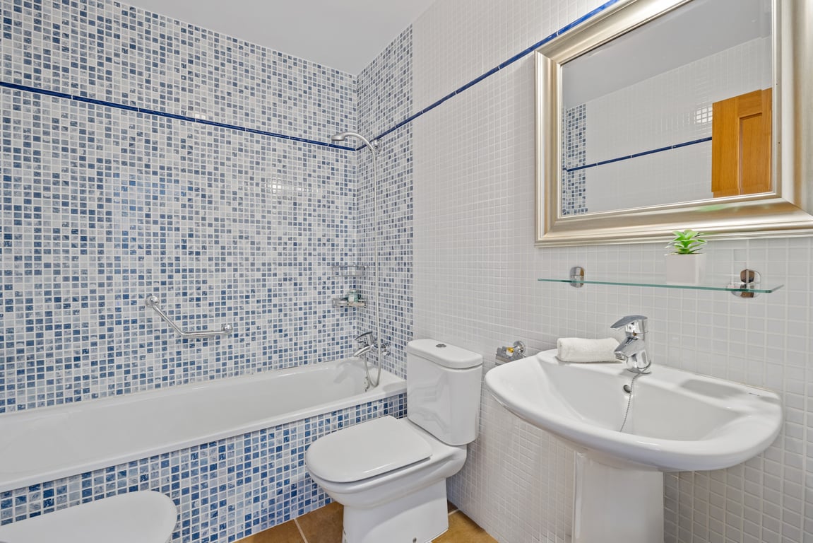 Cuarto de baño funcional con bañera integrada, paredes revestidas en mosaico azul y blanco, lavabo de pedestal y un amplio espejo con marco plateado.