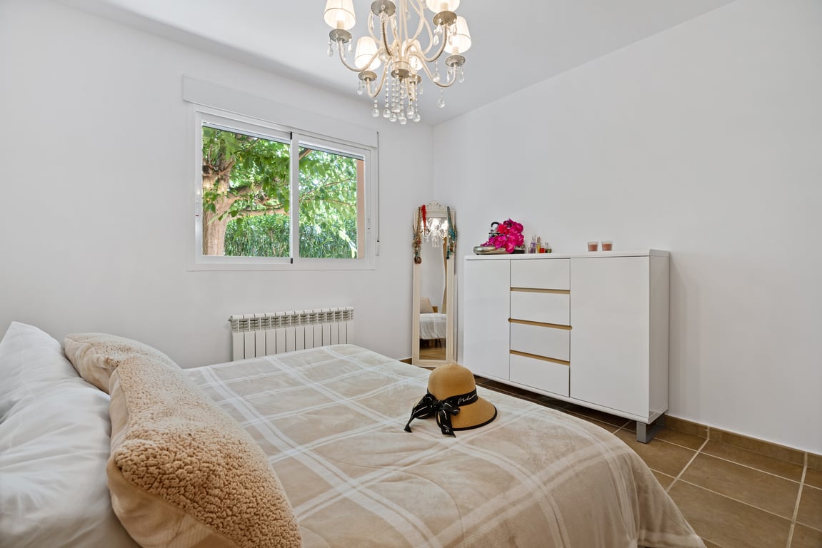 Dormitorio luminoso con suelos cerámicos, ventana de doble acristalamiento con vistas al jardín, radiador de calefacción central y una elegante lámpara de araña.