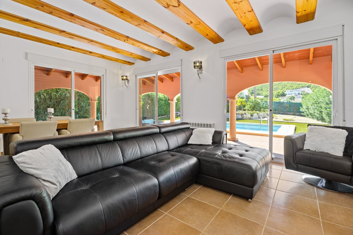 Salón luminoso con techos de vigas de madera, sofá de cuero negro y amplios ventanales correderos que conectan directamente con la naya y la zona de la piscina.