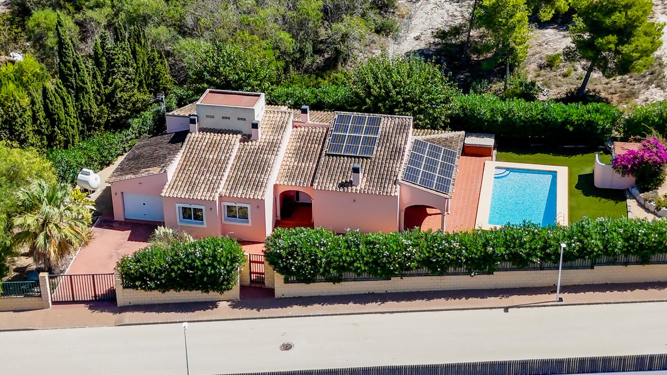 Vista aérea de la villa destacando la piscina privada, el tejado de teja con paneles solares y la parcela vallada con garaje y acceso pavimentado en Jávea.
