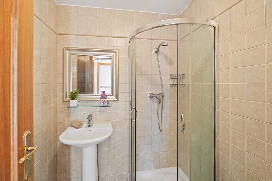Cuarto de baño con cabina de ducha de esquina acristalada, lavabo de pedestal blanco y paredes revestidas con azulejos en tonos crema.