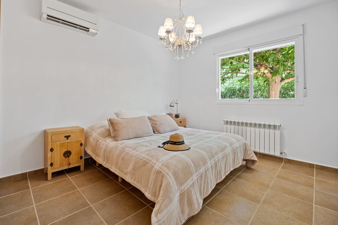 Dormitorio con suelos cerámicos, aire acondicionado split, radiador y ventana con vistas al jardín. Destaca su lámpara de araña y mesitas de noche de madera.
