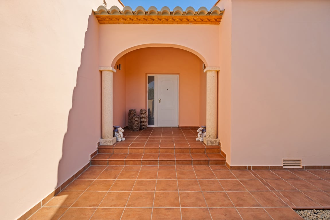 Entrada principal con porche arqueado sostenido por columnas de piedra natural y suelos de terracota. Puerta blanca con panel lateral de cristal.