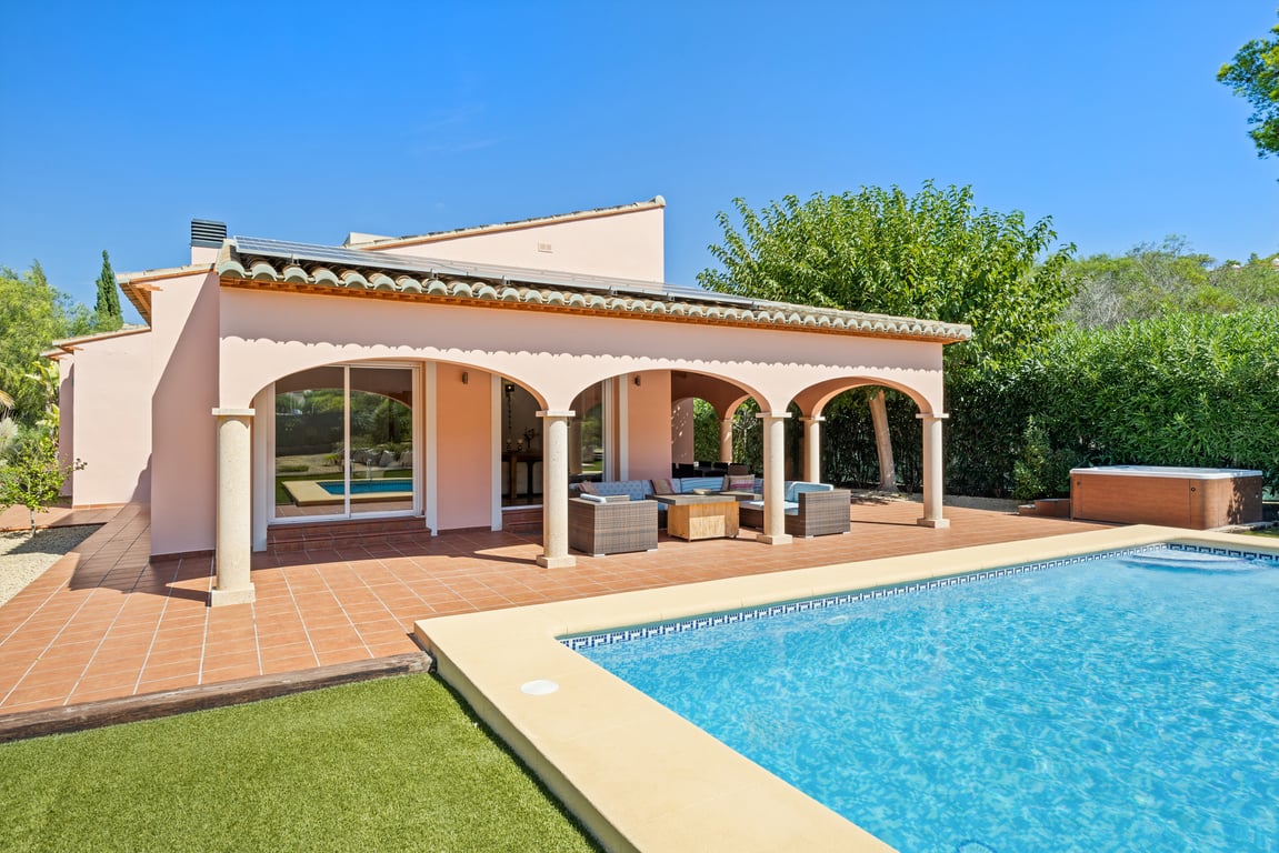 Exterior de la villa con naya arqueada, pilares de piedra y terraza de terracota frente a la piscina de mosaico, paneles solares y jacuzzi lateral.