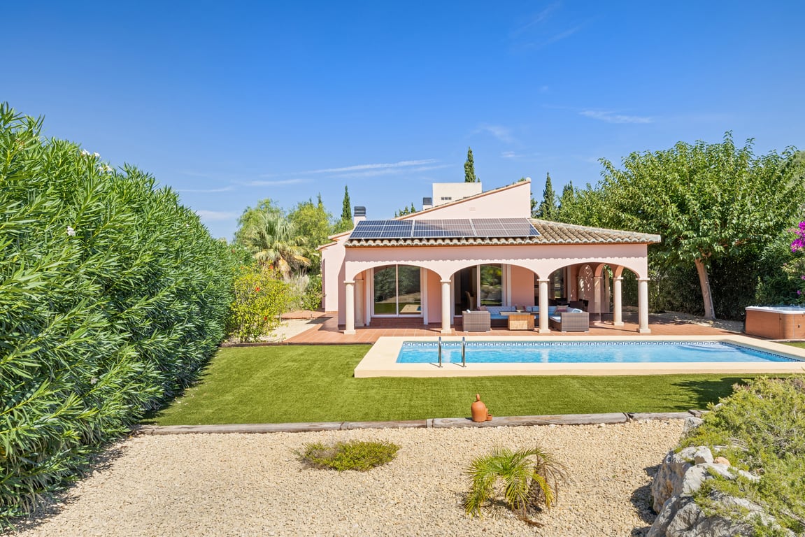 Exterior de la villa con piscina, amplia naya de arcos, paneles solares fotovoltaicos y jardín de bajo mantenimiento con césped artificial y setos maduros.
