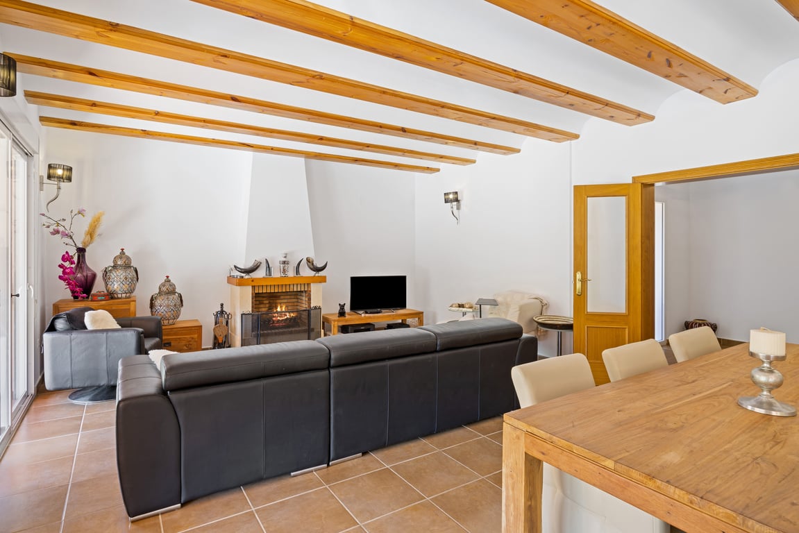 Salón comedor con vigas de madera a la vista, chimenea de obra y suelos cerámicos. Incluye zona de estar con sofá de cuero y mesa de comedor de madera maciza.