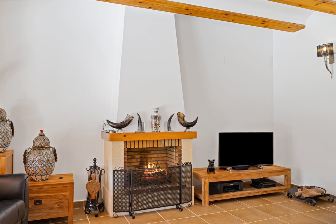 Salón con chimenea de obra, vigas de madera vista en el techo y suelos cerámicos. Incluye mobiliario de madera y detalles decorativos tradicionales.