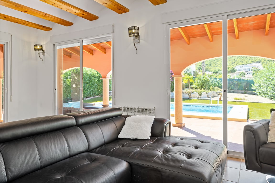 Salón con vigas de madera vistas y sofá de cuero negro, con acceso directo a la naya y vistas a la piscina y las montañas a través de grandes ventanales.