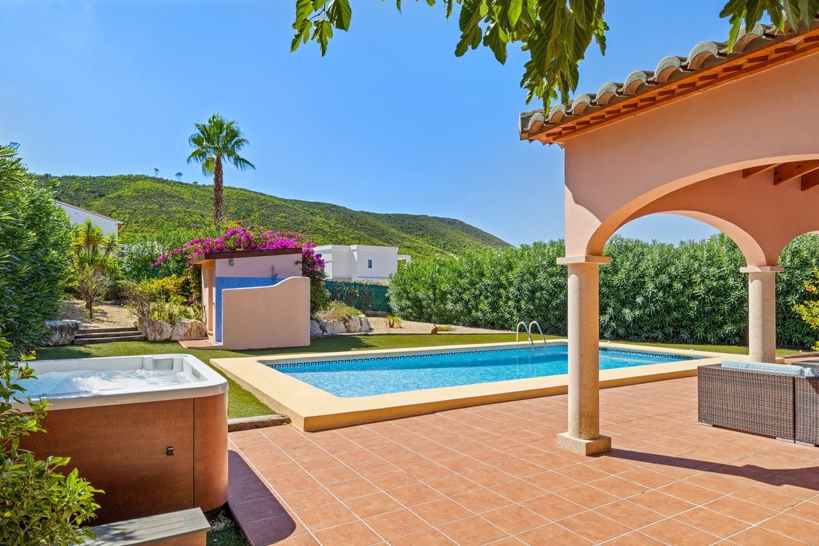 Terraza exterior con piscina de mosaico, jacuzzi independiente y naya con arcos, rodeada de jardín con césped artificial y vistas a la montaña.