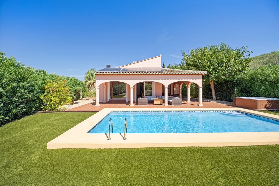Vista exterior de la piscina de mosaico y naya con arcos, rodeada de césped artificial, paneles solares en el tejado y un jacuzzi lateral.