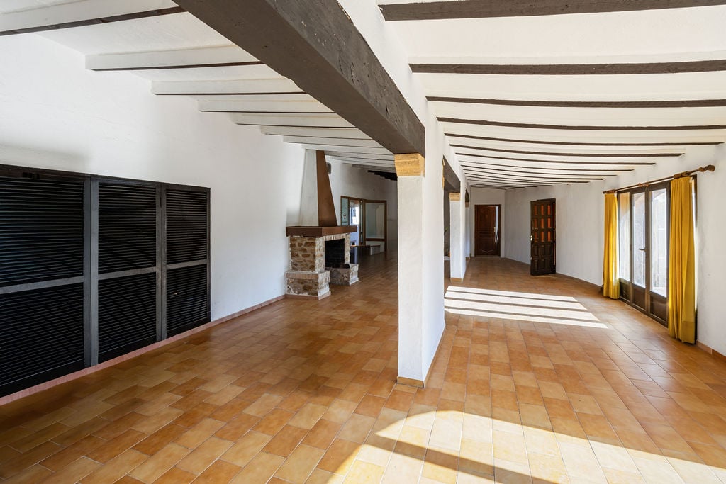Amplio salón diáfano con vigas de madera vista, suelos de terracota y una chimenea rústica de piedra. La estancia destaca por su luz natural y techos altos.
