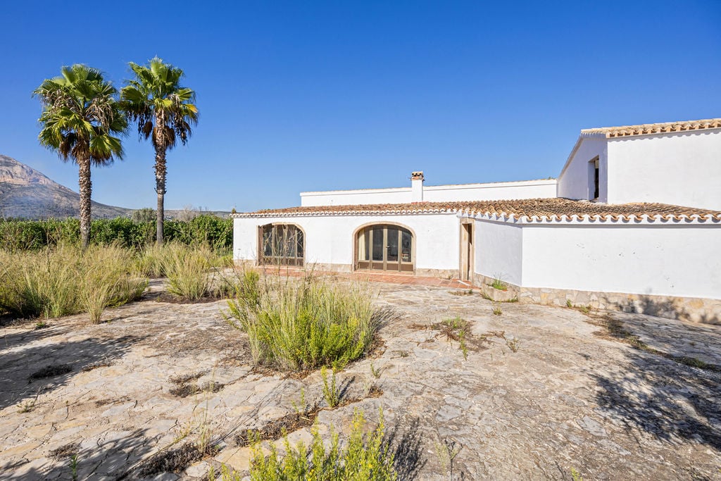 Exterior de estilo mediterráneo con fachada blanca, techos de teja y amplia terraza de piedra natural, flanqueada por palmeras y vistas al Montgó.
