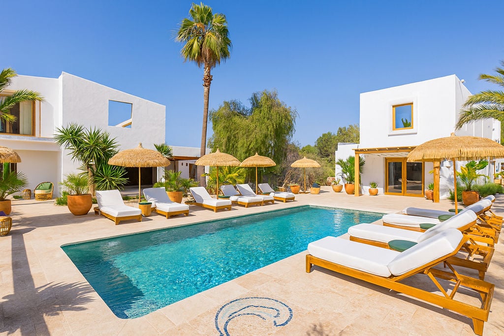 Piscina exterior rodeada de pavimentos de piedra clara, hamacas de madera con cojines blancos y sombrillas de brezo bajo una arquitectura mediterránea blanca.