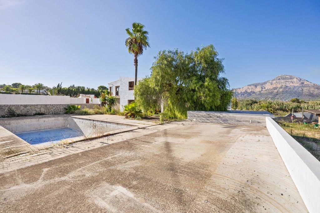 Terraza exterior de hormigón con vistas panorámicas al Montgó, piscina para rehabilitar y edificación de estilo mediterráneo rodeada de palmeras.