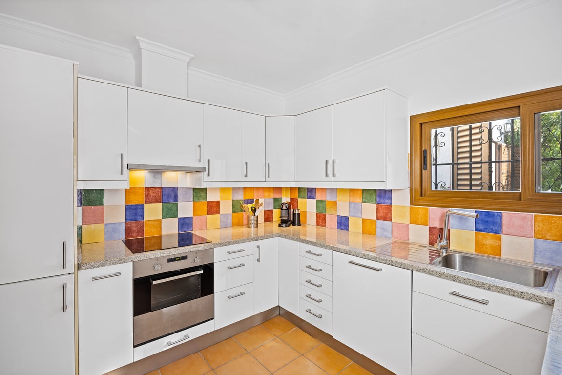 Cocina equipada con armarios blancos, encimera de granito y un frontal de azulejos cerámicos artesanales de colores. Incluye horno integrado y ventana con marco de madera.