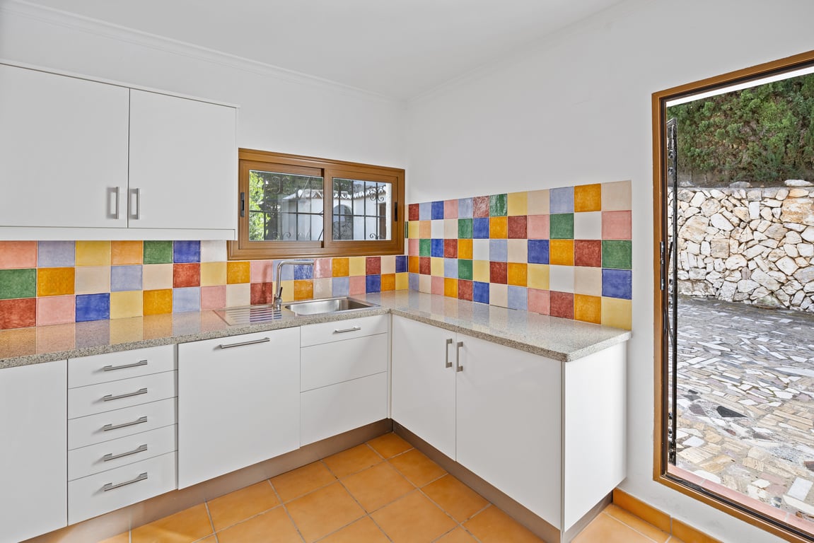 Cocina equipada con armarios blancos, encimera de granito y un llamativo frente de azulejos de colores. Dispone de suelo de barro y salida directa al patio lateral.