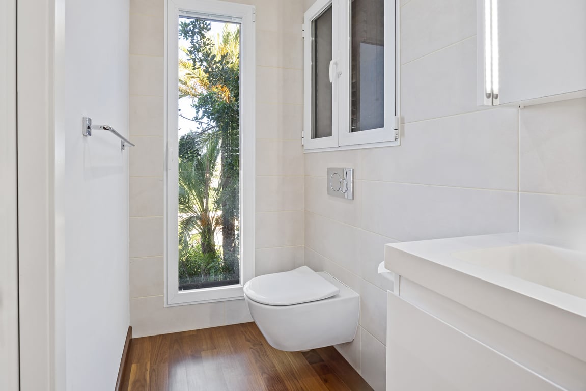 Cuarto de baño reformado con inodoro suspendido, suelo de madera y ventana vertical con vistas al jardín mediterráneo.