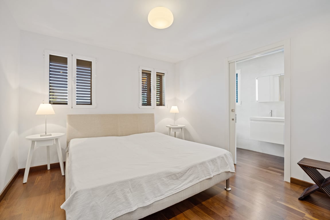 Dormitorio reformado con suelo de madera oscura, cama doble y acceso directo a un baño en suite moderno mediante una puerta corredera blanca.