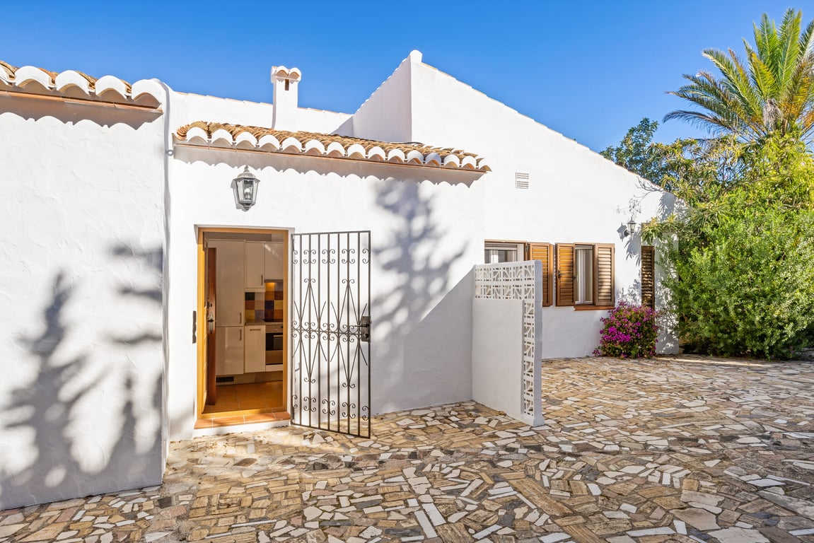 Fachada mediterránea con paredes blancas, pavimentación de piedra irregular y acceso secundario a la cocina con reja de hierro forjado y contraventanas de madera tradicionales.