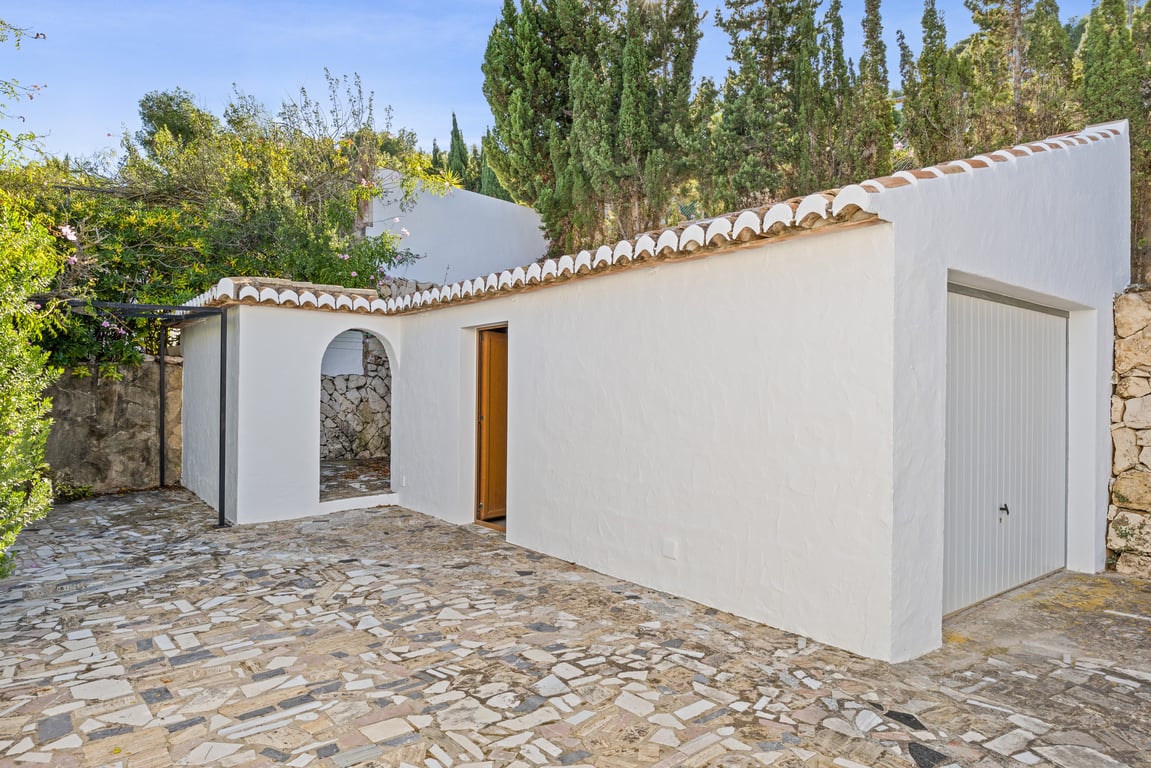 Garaje independiente y zona de servicio con fachada blanca, tejado de teja cerámica y suelo de mosaico de piedra irregular rodeado de vegetación mediterránea.