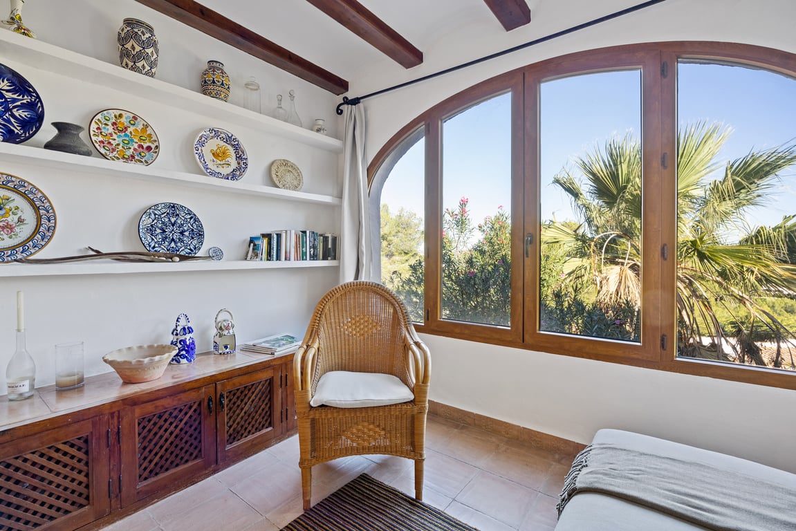 Naya acristalada con vigas de madera, estanterías de obra con cerámica tradicional y un gran ventanal arqueado con vistas al jardín mediterráneo.