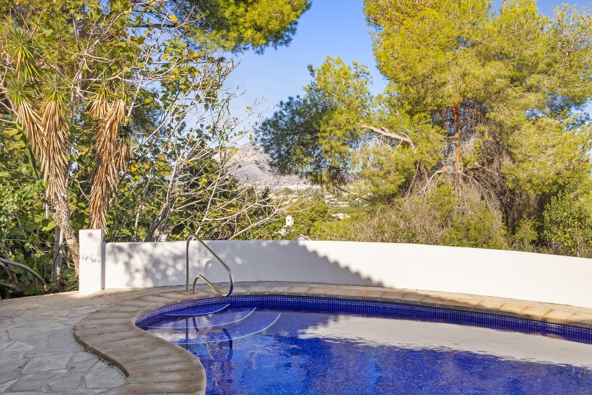 Piscina de gresite azul con escalera metálica, rodeada de una terraza de piedra natural y vegetación mediterránea con vistas lejanas al macizo del Montgó.
