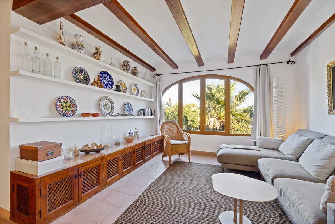 Salón con vigas de madera, estanterías de obra con cerámica tradicional y gran ventanal arqueado con vistas al jardín mediterráneo.