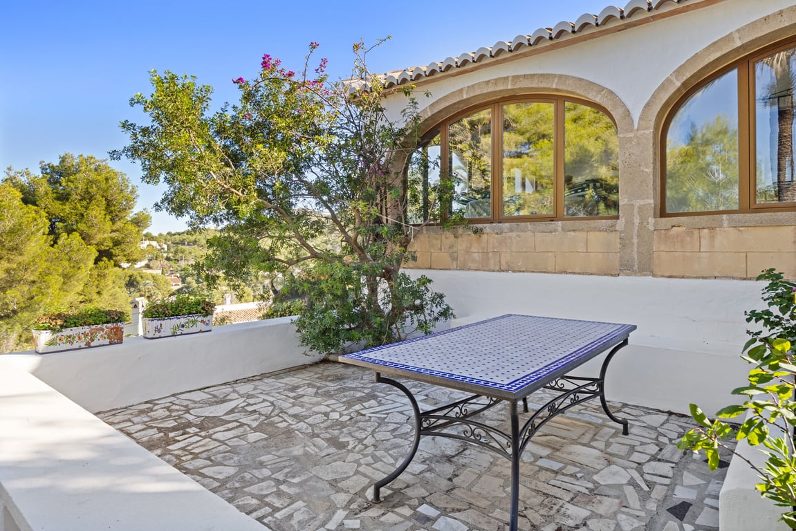 Terraza exterior con suelo de piedra irregular, mesa de azulejos artesanales y arcos de piedra Tosca que enmarcan la naya acristalada bajo el sol mediterráneo.