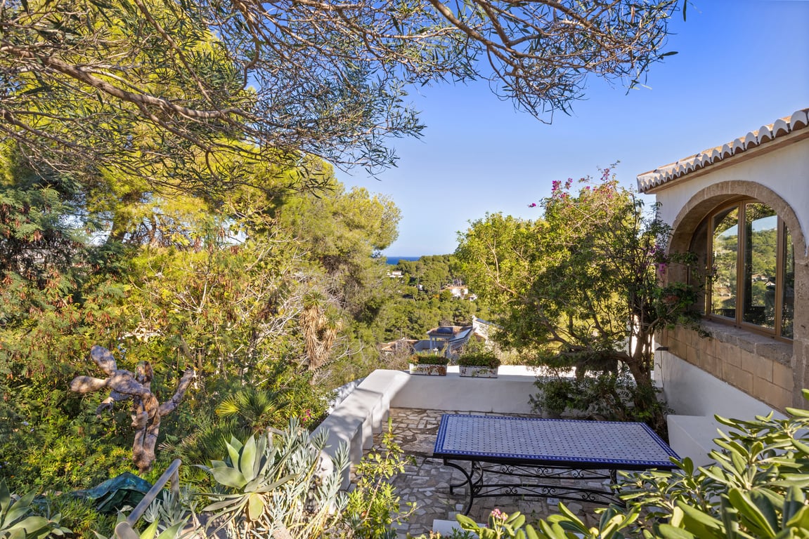 Terraza exterior con suelo de piedra natural y mesa de mosaico, rodeada de vegetación mediterránea y vistas al mar enmarcadas por pinos y arquitectura tradicional.